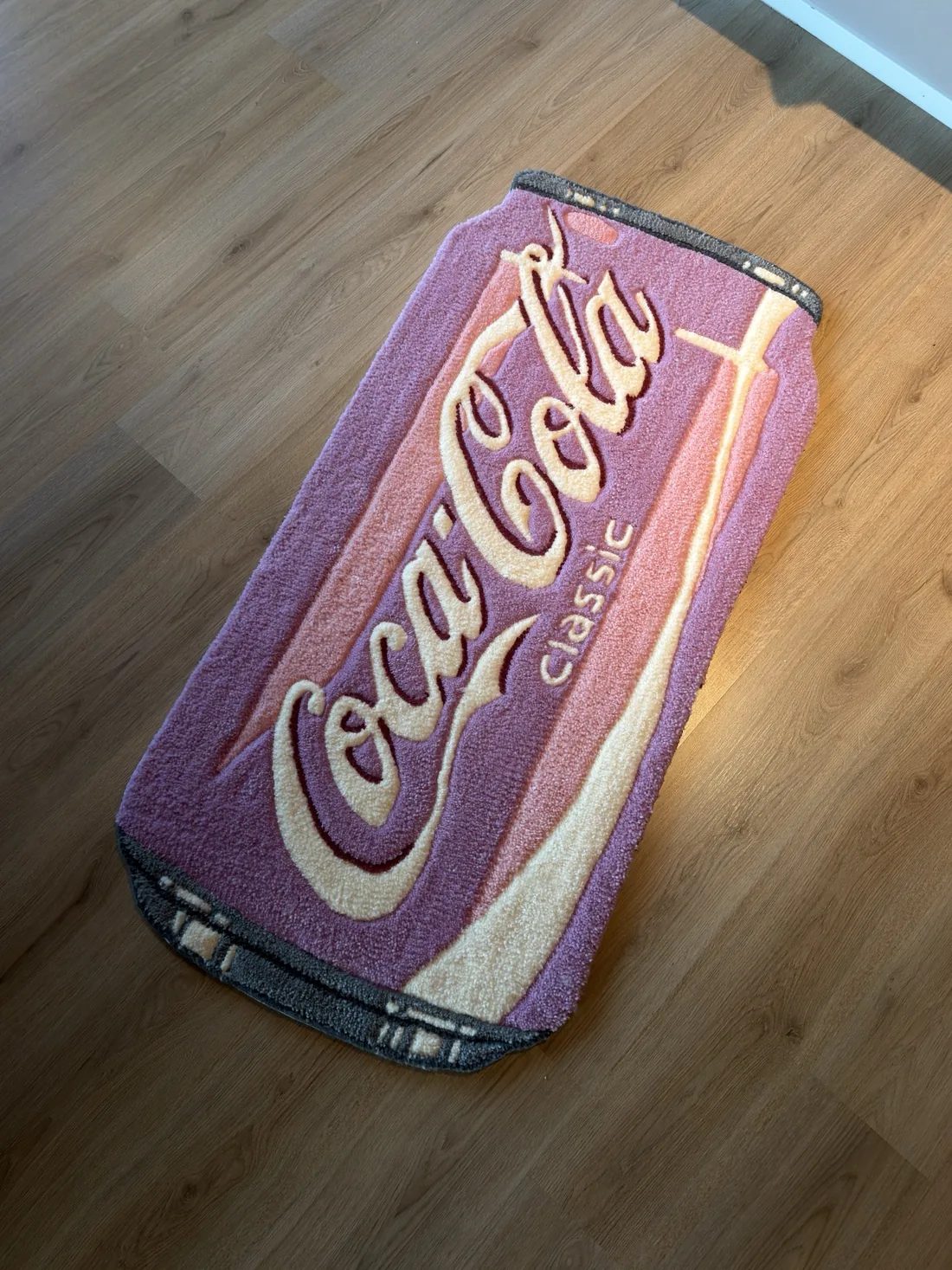 Teppe formet som en brusboks med Coca-Cola-logo, liggende på et tregulv